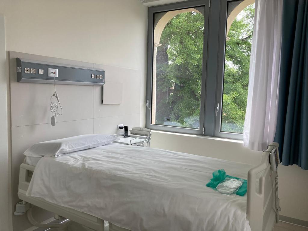 Degenza - Camera di degenza singola luminosa con letto ospedaliero regolabile, grande finestra con vista su alberi, tenda azzurra e attrezzatura medica a parete